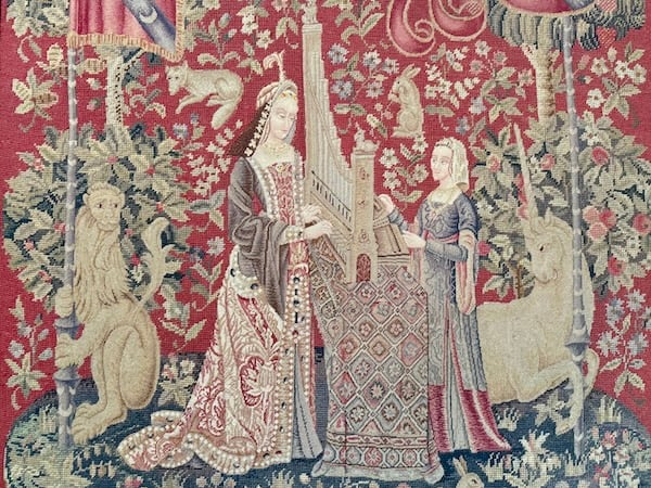 Woven tapestry showing a noble medieval lady playing a portable organ accompanied by a maidservant pumping the bellows. The women wear colorful gowns and are flanked by a lion and a unicorn holing flagpoles. They stand on a round blue island against a red background covered in woven flowers, foliage, and animals.