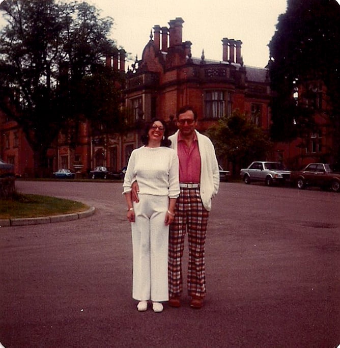 Chic woman in all white and man in plaid pants, white belt, pink shirt, and white cardigan, posing outside on London street