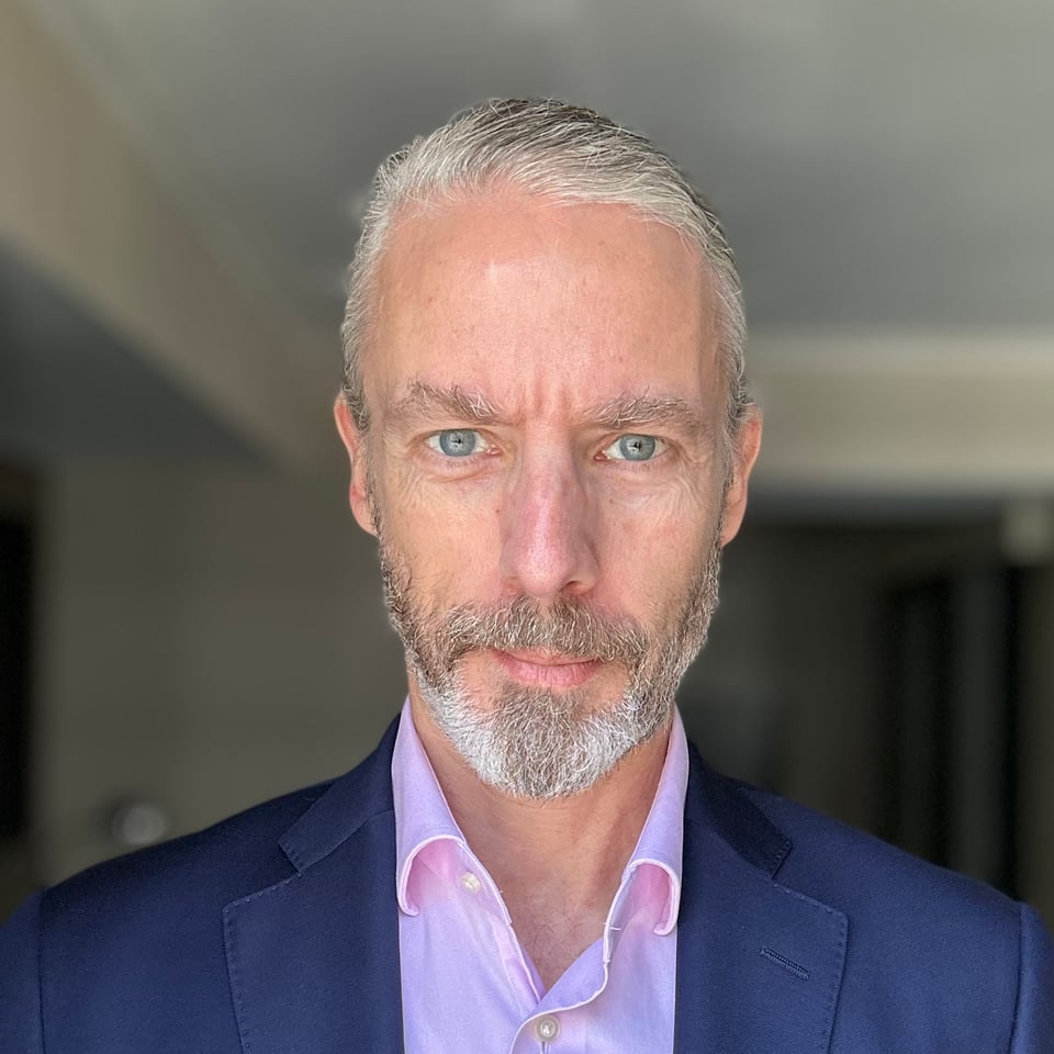 Close-up photo of a white man in a blue suit & pink dress shirt. His long hair is pulled back, and he clearly hasn’t slept much.