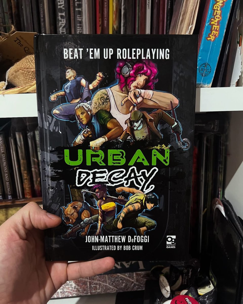 A hand holding a hardcover book. The cover reads Urban Decay, and depicts characters of a beat'em up arcade game.