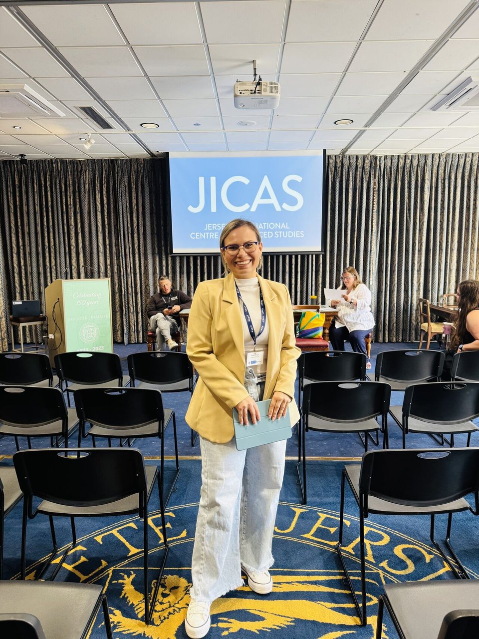 Thais Franken standing in a conference room, in front of the screen that highlights the name of the conference: JICAS.