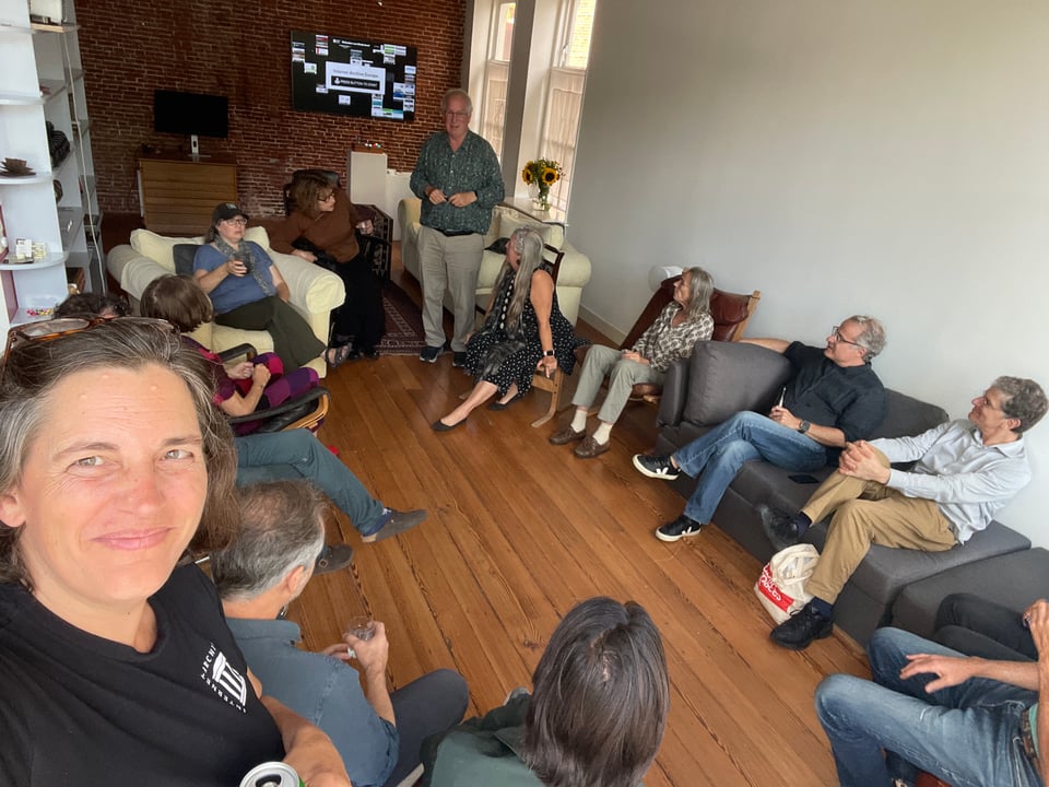 A group of people sit in a circle as a man stands and addresses them all.