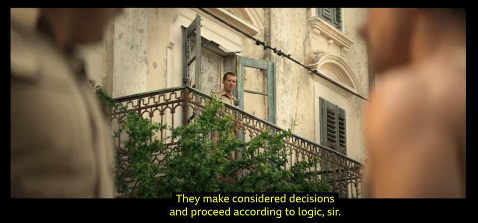 Screenshot from the TV series, SAS Rogue Heroes. In the background an army officer is standing on the balcony of an old building in a Sicilian town. The officer is looking down on a conversation between two soldiers who appear blurred in the foreground. We also see some subtitle text which says, "They make considered decisions and proceed according to logic, sir."