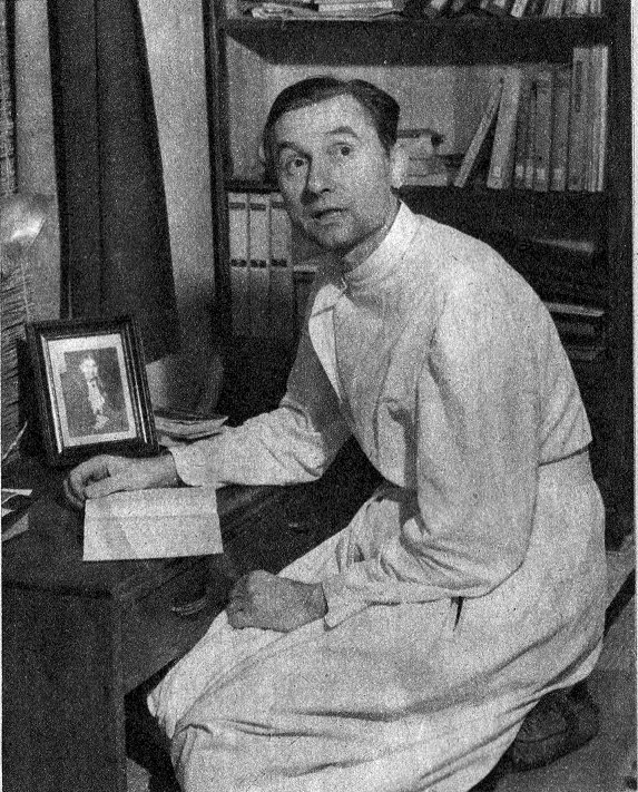 A Jesuit Priest in a white smock kneels at a desk, reading a passage from a book