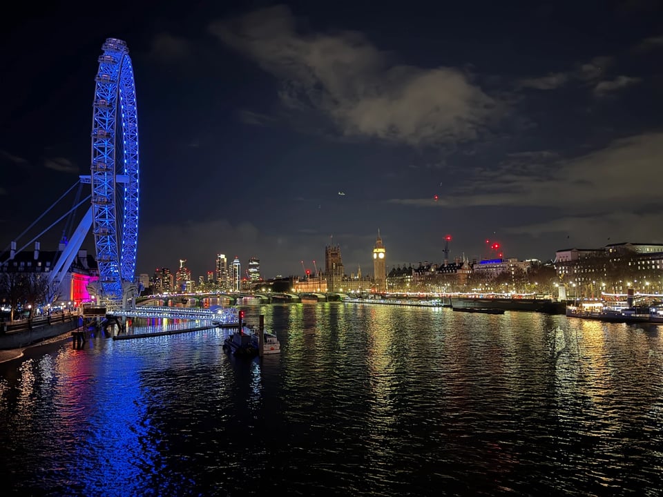 London skyline at night over the Thames. The London Eye lit in blue is on the left. The Houses of Parliament and the Elizabeth Tower which holds Big Ben is on the right.