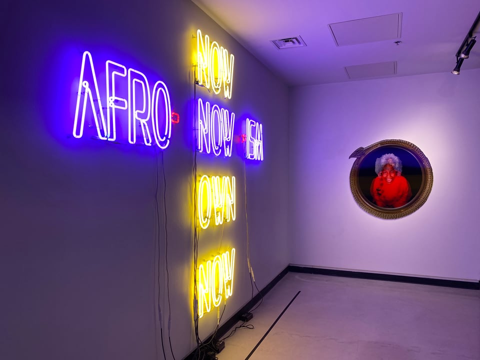 A gallery wall with an acrostic-like neon sign that says Afro-now-ism across and Now Now Own Now down. On another wall is a large, circular lenticular image of a Black woman leaning forward and laughing.