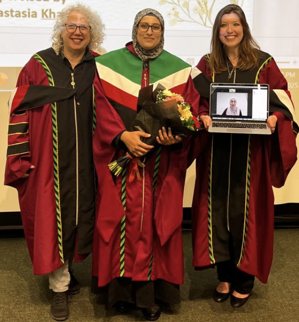 Three women standing in red academic regalia; the woman on the right holds a laptop with an image of a fourth woman via videoconference