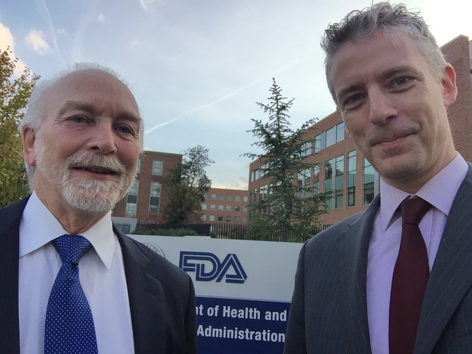 Photo of 2 white men in suits and ties in front of FDA headquarters. The man on the right is in his mid-40s and the one on the left is in his mid-60s.