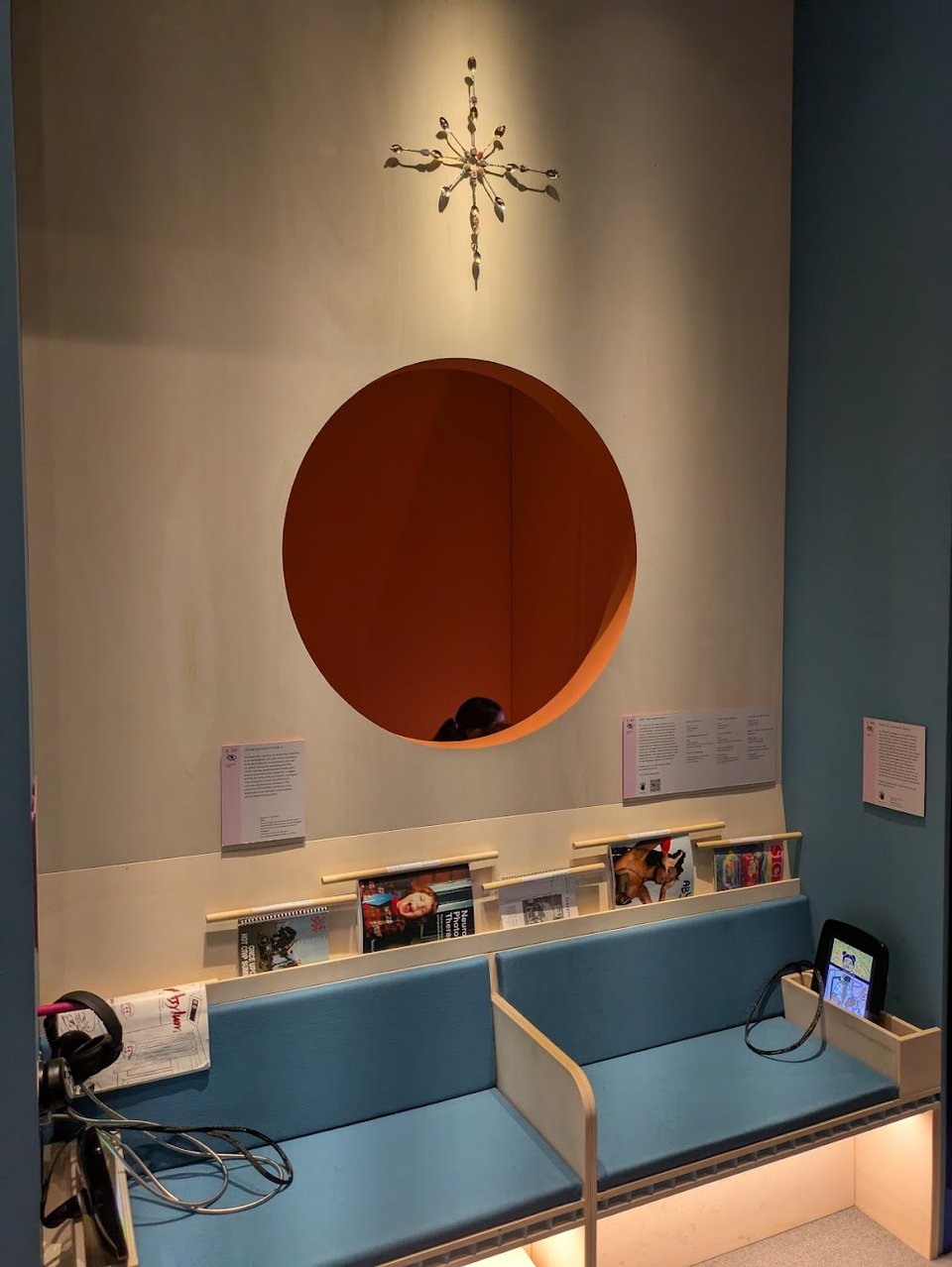 A seating area with magazines and tablets is attached to a wall. Above the seats, a large hole in the wall makes it possible to see people on the other side. There is the top of someone's head visible through the hole.