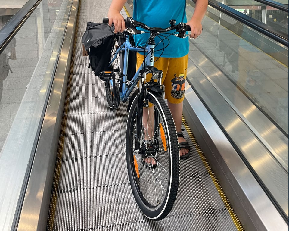 A boy seen from the waist down stands on a ramp escalator going down from a bike store holding his brand new light-blue 26" wheel bicycle.
