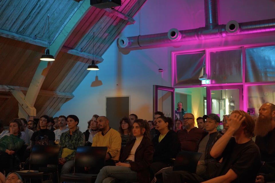 an audience, backlit in purple and blue, listens to an off-frame presenter