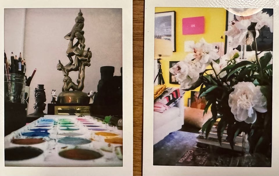 Two Instax photos: left is a tray of watercolor pigments in the foreground, leading back to an award trophy for the cartoonist Patrick McDonnell, flanked by his drawing tools; right is a photo of some white flowers on the edge of going off, on a coffee table in front of a yellow wall