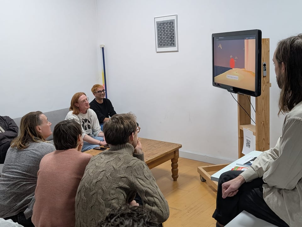 A group of people in couches are seated around a TV, listening to a seated speaker in a white t-shirt. The screen shows a low-poly videogame scene on a beach. The sky is purple.