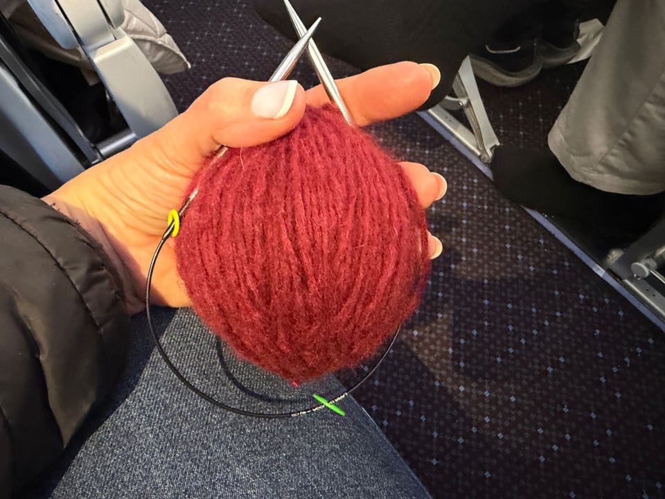 A woman wearing a pair of jeans sits in an airplane. She is holding a ball of dark red wool and a pair of knitting needles attached "on the round".