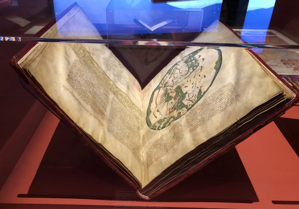 A medieval manuscript in a museum case. The two two-thirds of the right-hand page have a circular world map on it: green sea, white land, and a few geographical features.