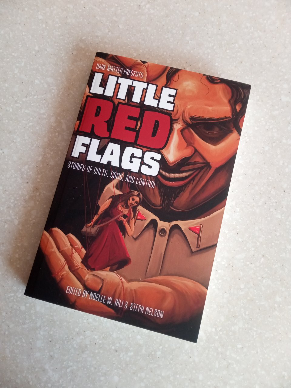 A copy of LITTLE RED FLAGS: STORIES OF CULTS, CONS, AND CONTROL edited by Noelle W. Ihli and Steph Nelson. Cover features a sinister-looking man holding a puppet of a woman in a red dress on his palm, while his other hand holds the strings. MWA-HA-HA!