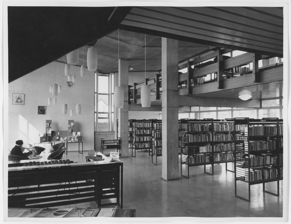 An internal photo of the original building from the south west corner. Light is coming in from the eastern curtain wall to illuminate metal bookshelves. H shaped beams hold up the student gallery. Oval frosted glass lights hang over an issuing desk, and wire-frame metal armchairs are set around tables. An older lady in a hat is sat in one, reading a newspaper.