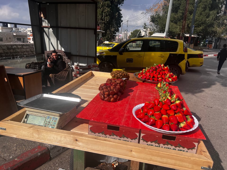 With taxis in the background, a table holding a beautiful display of two platters of big strawberries piled high, and a few bags of brown chestnuts. A scale sits on the table as well.  