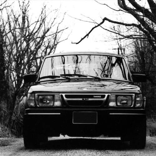 A black and white photo of a car taken from directly in front of it. It's close enough that it takes up most of the frame. The car is a Saab 900 S, a small, boxy kind of car. There is a figure in the driver's seat but the reflections of the trees in the windshield obscure their identity. (It's musician Will Oldham of Bonnie "Prince" Billy.) The dirt road and leafless branches on either side suggest a country road. The words "SLINT" AND "TWEEZ" are on the grille of the car.