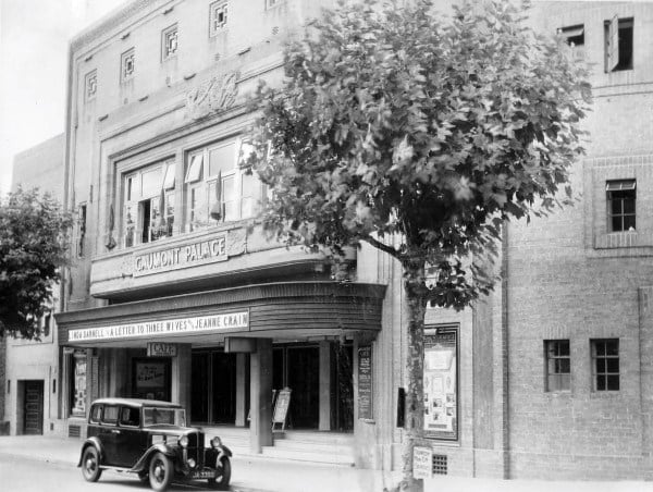 A 1930s honey-coloured brick cinema with Deco detail. The main front is a giant squared arch, with rib detail. A second square arch surrounds large windows set behind a curved balcony. Below that is the Gaumont Palace sign and the doors. An early car, maybe an Austin?, is parked in front.