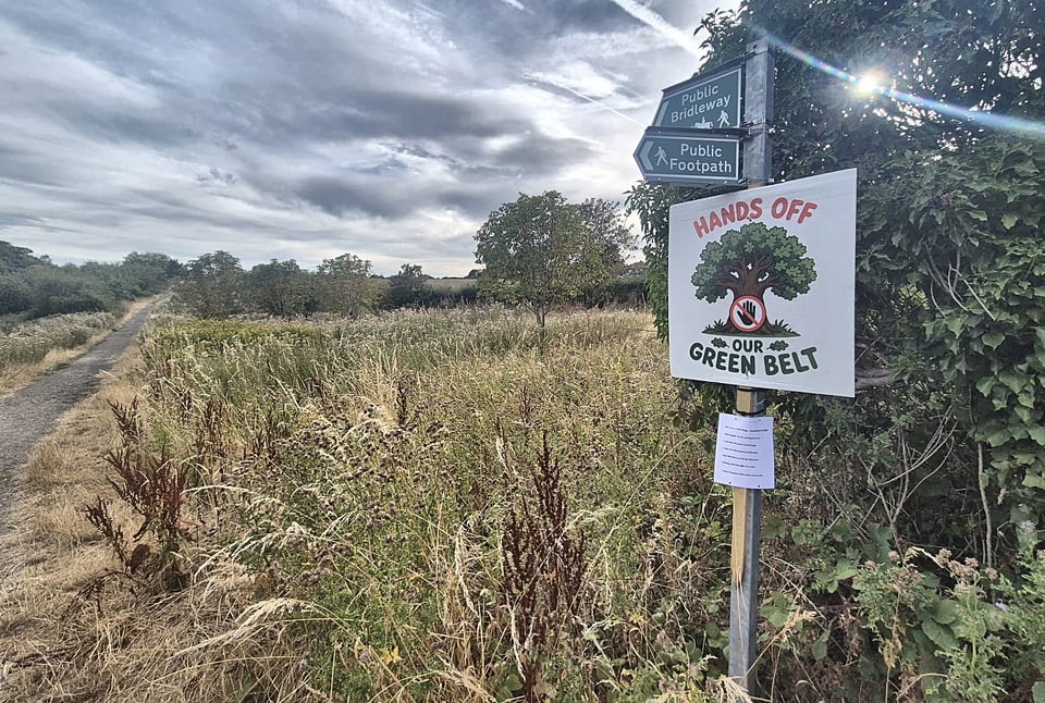 A public bridleway and public footpath fingerpost beside a track crossing rough grassland, with a campaign sign fixed below reading Hands Off Our Green Belt.