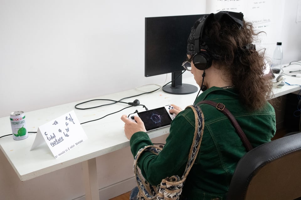 A person plays a game on a handheld device next a sign that reads "c o a l e s c e n t tidal rapture"