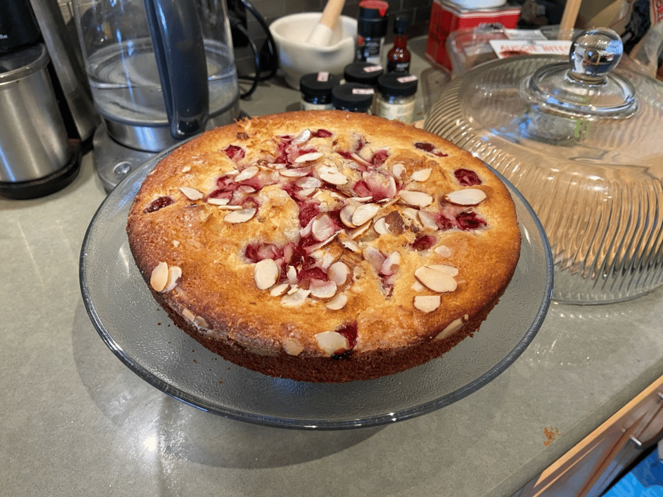 A simple cake with red berries in it and no icing sits on a glass cake stand.