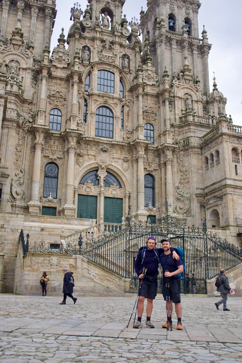 Two bearded pilgrims with trekking poles and full packs embracing in the Praza do Obradoiro, the ornate Baroque facade of the Cathedral of Santiago de Compostela towering behind them at journey's end
