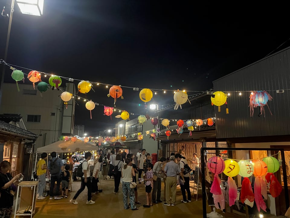 Image of lanterns strung across a narrow busy street where Tanabata festival goers are gathered to watch a live jazz band.