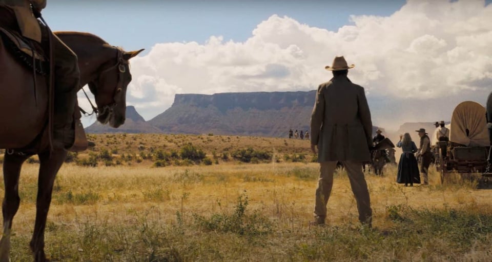 A still from Horizon: An American Saga - Chapter 1 of the back of a cowboy looking into the plains. There's a horse to the left and a wagon to the right.