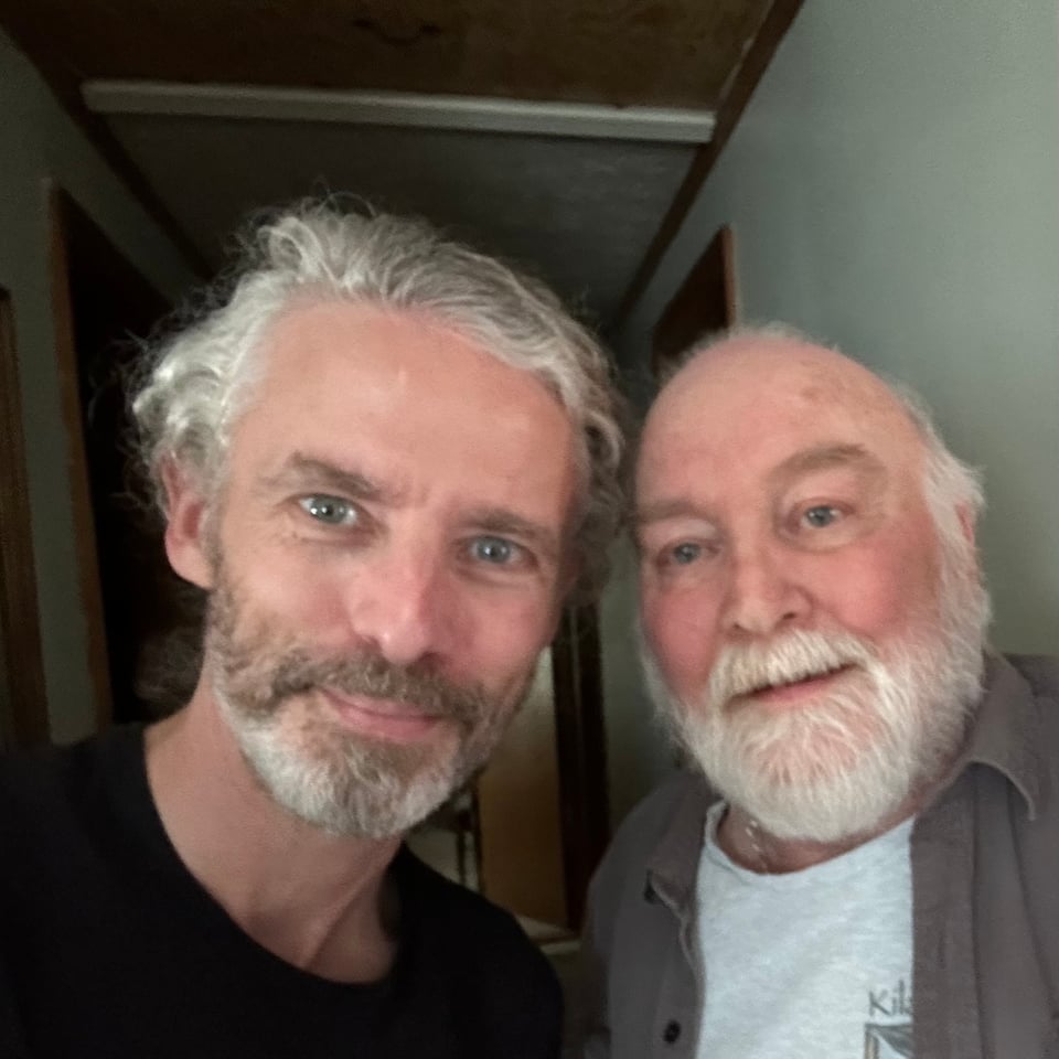 Photo of 2 white men in a house. Both are dressed casually, smiling for the camera. The man on the left is in his early 50s and the man on his right is in his early 70s. The man on the right is dying of a brain tumor, and his face is puffy from the steroids that are part of his treatment.