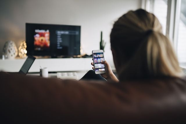 generic but probably not yet synthetic image of a person looking at their phone while sitting in front of a television