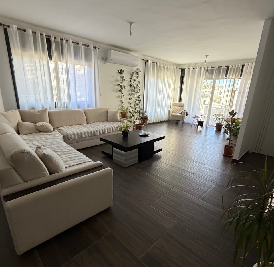 A living room with dark wood floors, sheer curtains, sofa, chair, coffee table and plants