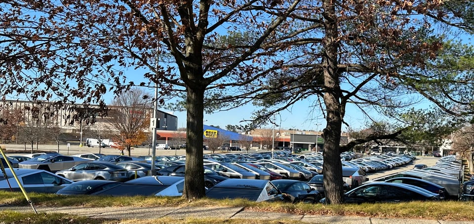Photo of parking lot that serves as storage for a Tesla dealership; not taken at a great angle, but the viewer can see there are DOZENS of Cybertrucks