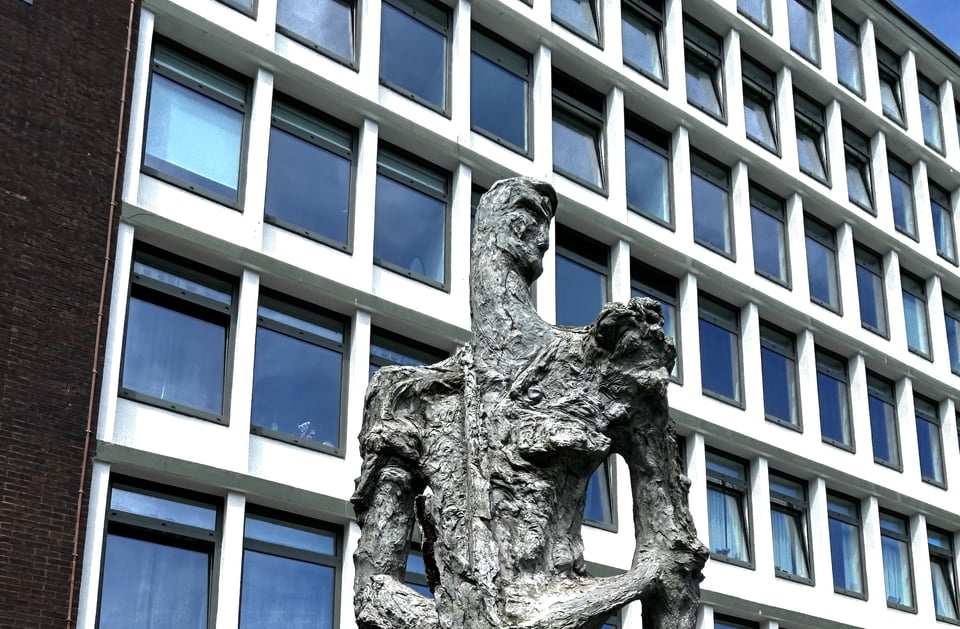 Close up of the upper half of the agriculture sculpture. It's a abstracted male figure holding a scythe blade down. His body is half melted and distorted like a Giocometti figure.