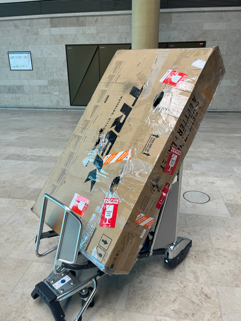 A large cardboard box on an airport luggage trolley covered in holes patched with tape and numerous "Fragile" stickers