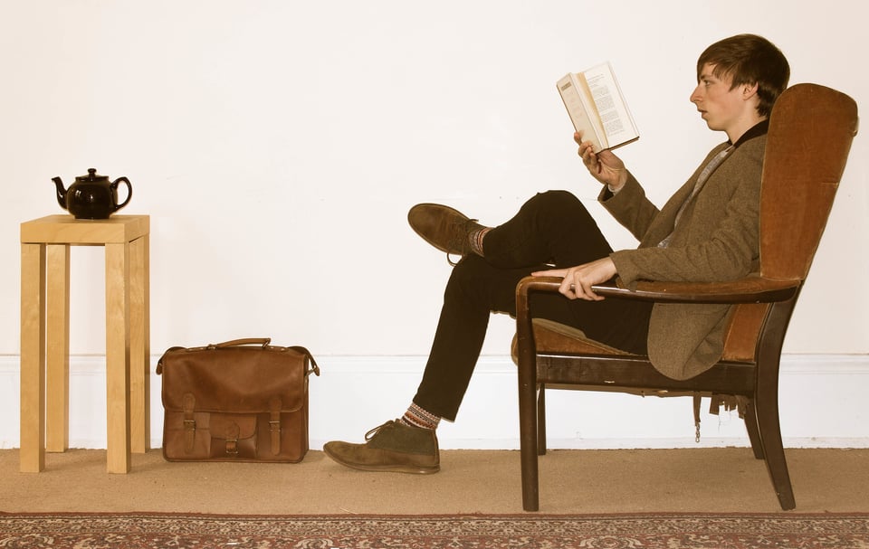 A person with brown hair and a pointy nose wearing a natty jacket, dark pants and cool brown shoes, sitting in an armchair reading a book, with a satchel and a table containing a teapot nearby. VERY HIPSTER.