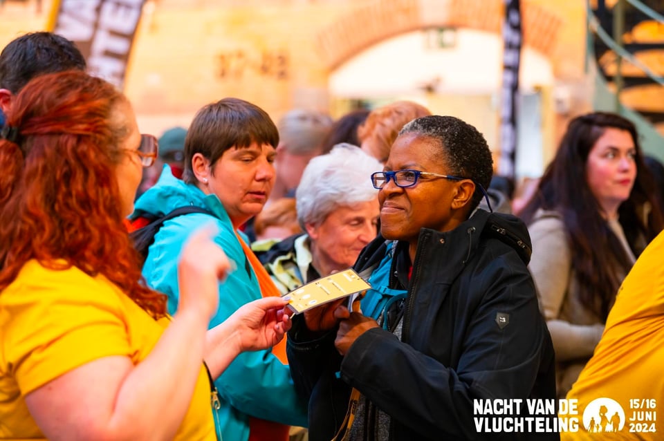five women of various ages and ethnicities; a white woman in a yellow t-shirt hands a tag to a black woman in a black jacket and glasses