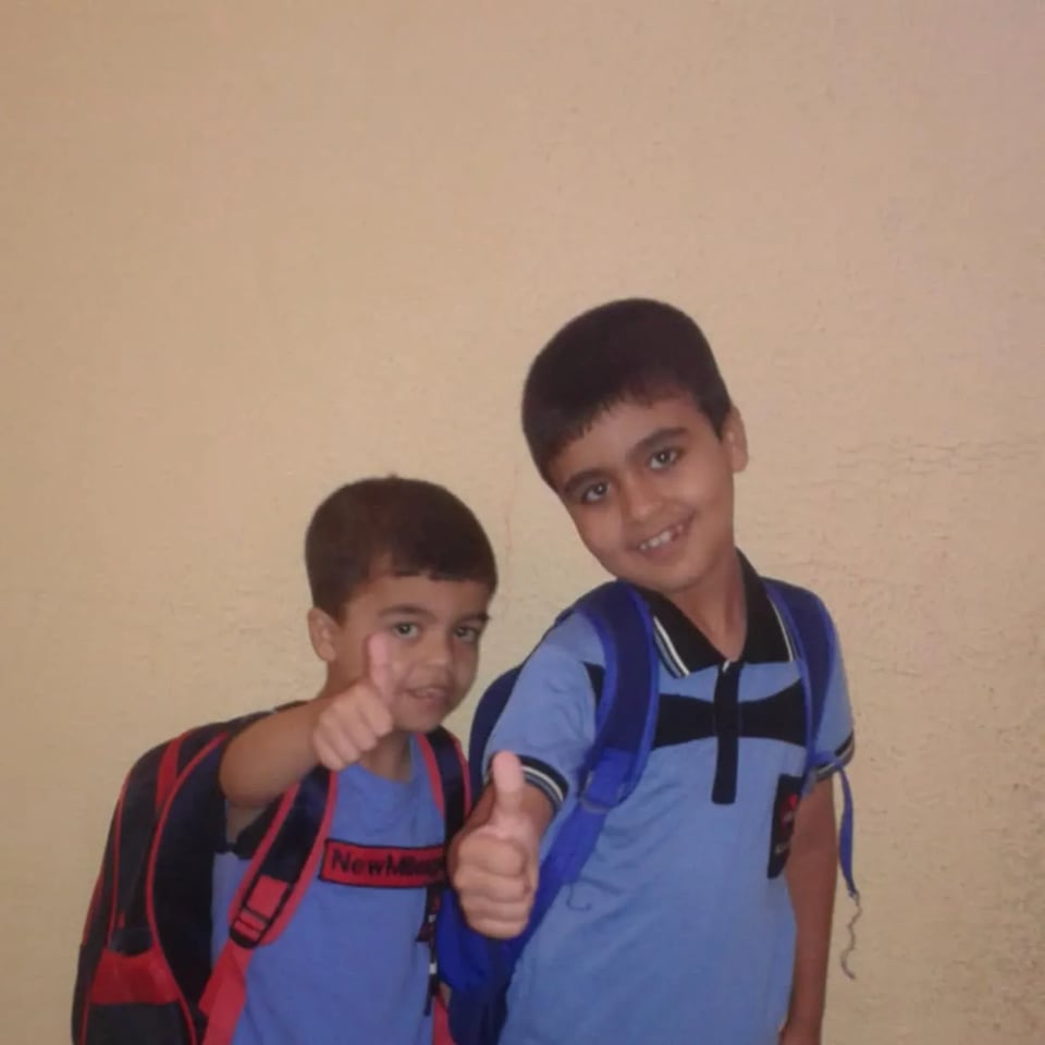 Two young children stand side by side against a plain light-colored wall, both wearing backpacks and blue shirts. They are smiling at the camera and giving thumbs-up gestures, suggesting excitement or confidence, as if ready for school.