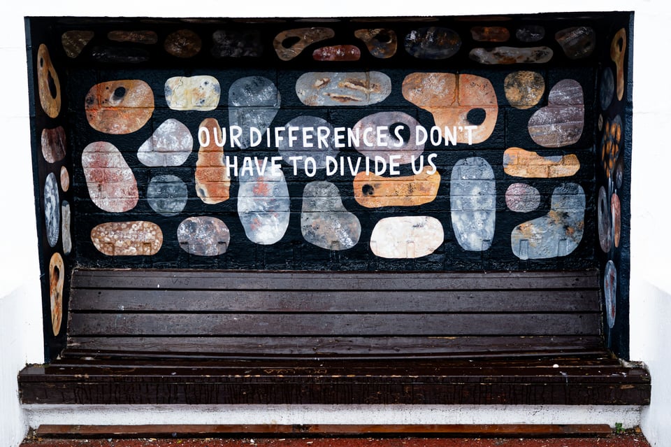 A wall over a bench with different oval shapes with different colors and designs. Over the shapes are the words "Our differences don't have to divide us"