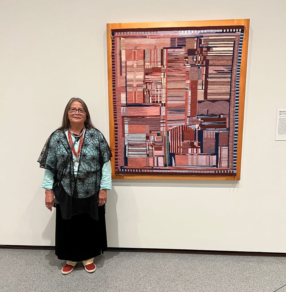 Photograph of Dine (Navajo) weaver Marilou Schultz standing next to one of her creations, the 1994 work “Replica of a Chip”. Click image to view source.