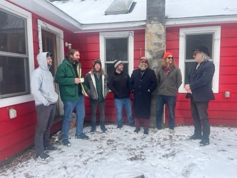 Seven men in winter coats stand outside in a open semi-circle in a snowy yard outside a red house.
