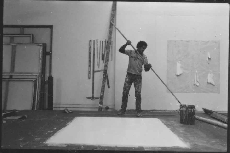 A black-and-white photo of the artist Jack Whitten in his studio. A large canvas is laid in front of him on the floor and he appears to be dipping a long pole into a paint bucket or perhaps in a paint tray behind the pail.