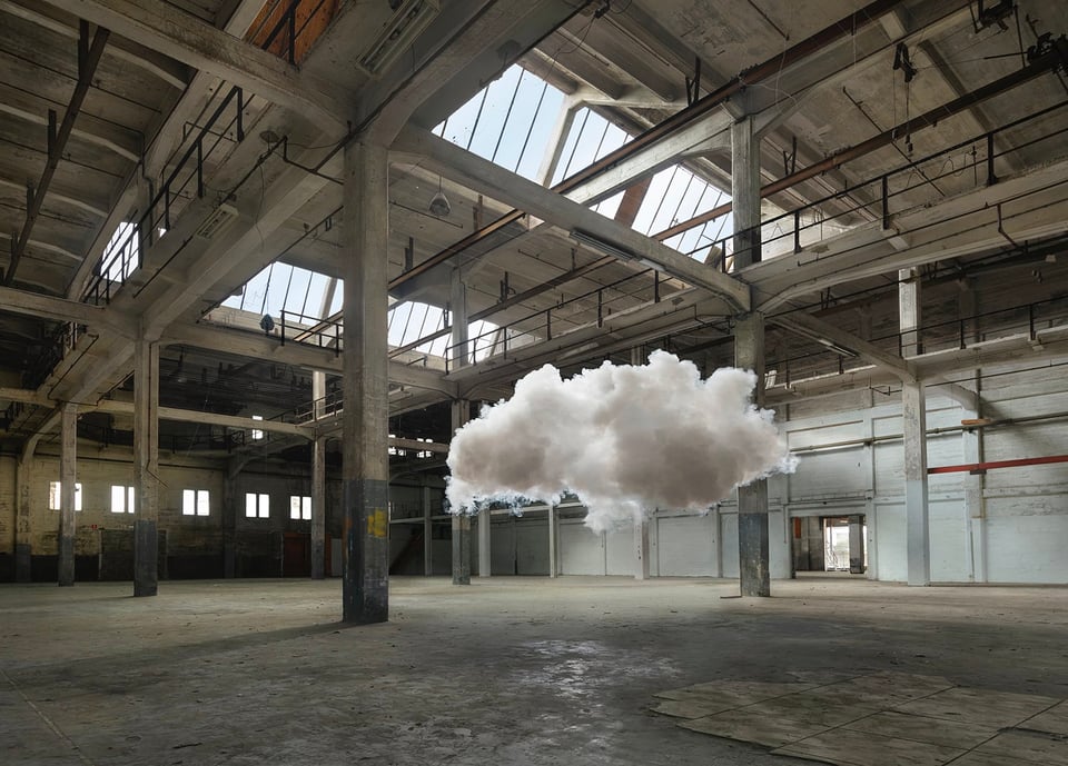 A white puffy cloud, perhaps 15 feet long and 8 feet high, floating inside an industrial building that looks like an empty factory floor. Sunlight comes in from a few windows in the ceiling