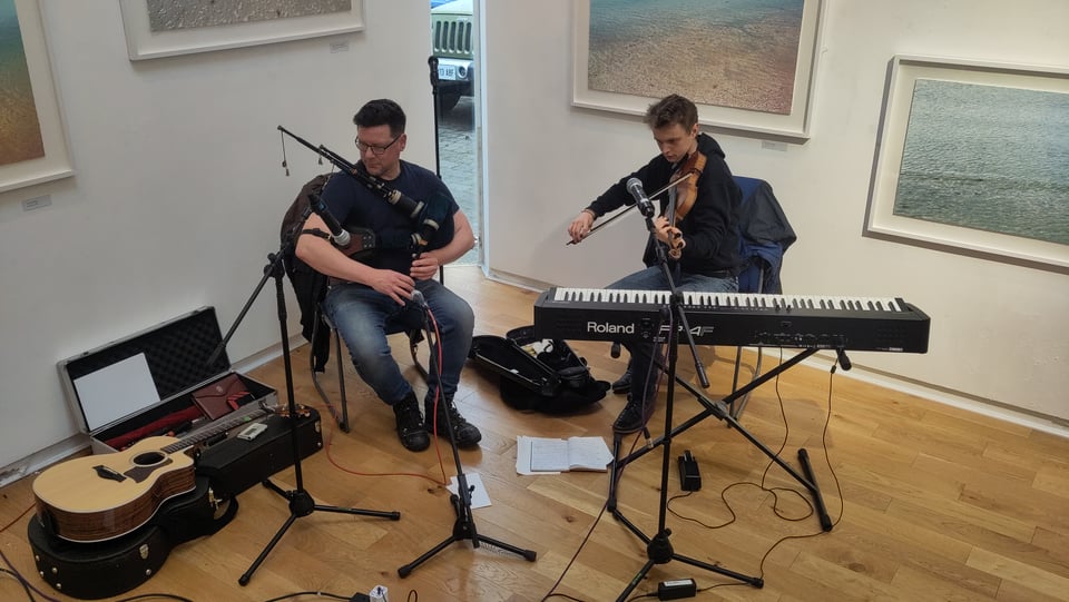 Donald WG Lindsay and Roo Geddes, seated in the rear corner of the gallery at An Lanntair in Stornoway, behind electric piano and microphones, and beneath print works showing sea shore scenes, playing the smallpipes and fiddle.