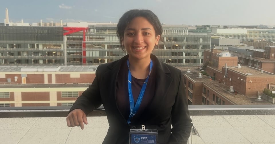 A JMC student in a black blazer and blue lanyard stands on a rooftop, smiling toward the camera. Behind her, modern glass buildings and brick structures form an urban skyline under daylight.