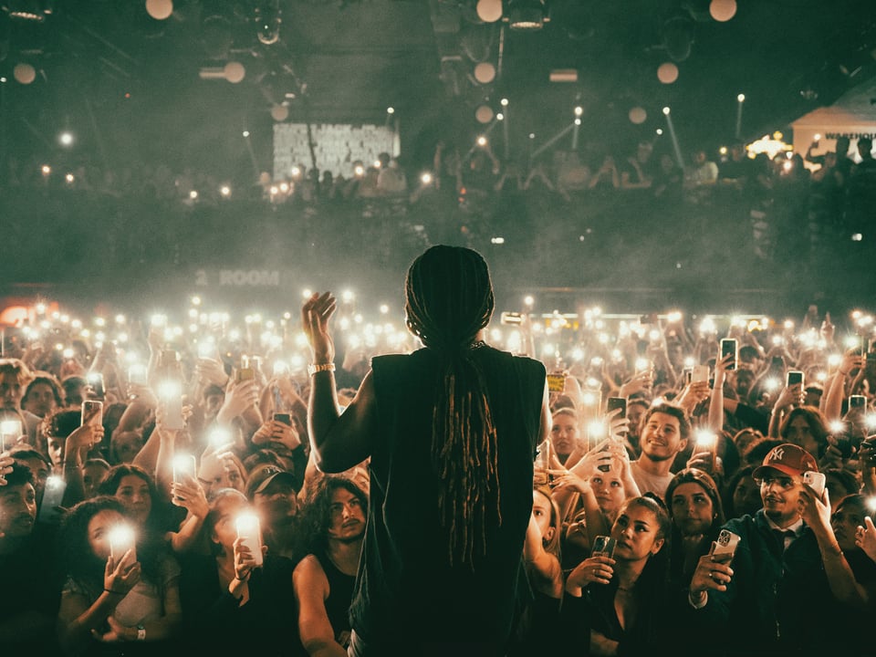 A performer on stage is facing away from the camera, silhouetted by the audience members shining their cellphone flashlights toward the stage.