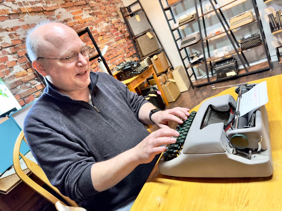 Glenn Fleishman sits in a chair at a table typing on a manual typewriter. The photo is at a slight angle. A brick wall is behind him and a wall of typewriters on shelving is at his left (upper-right of photo)