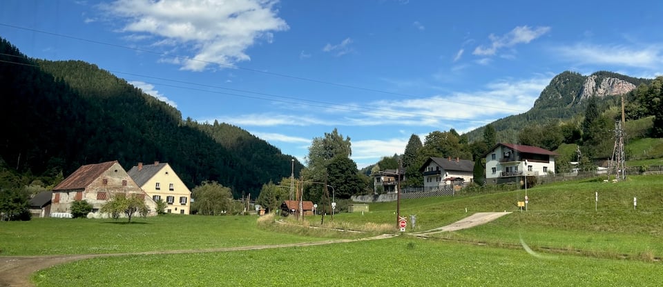 Some big hills, some wooden buildings, very, like, middle-European hilly trees and sky sort of thing