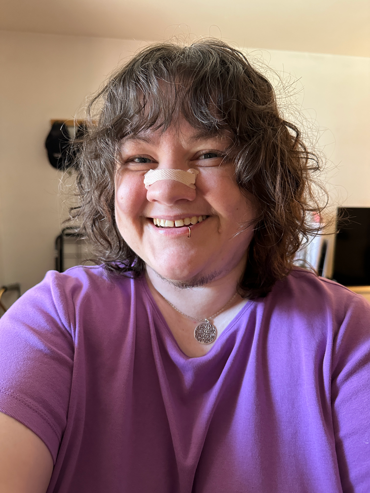Photo of Jessi with a shaggy mullet that emphasizes their brown waves / curls. They're sporting a huge grin, and they're wearing a purple shirt, a nose strip, a lip ring, and a Celtic knot necklace.